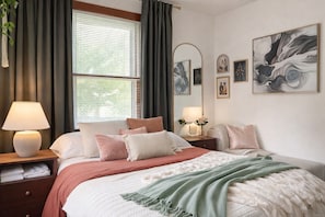 Un dormitorio con cama, mesitas de noche, lámpara, ventana con cortinas y decoración en la pared.