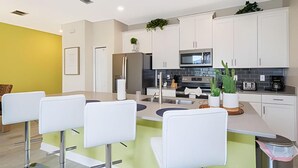 A modern kitchen with white cabinets, a stainless steel refrigerator, a microwave, and a sink. There are bar stools and a potted plant on the counter.
