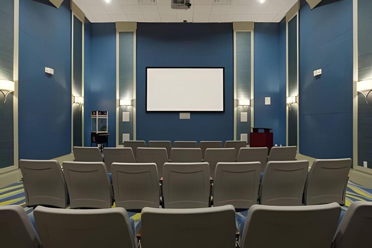 Konferenssihuone, jossa on riveissä tuoleja, ja niiden edessä suuri näyttö. Seinät ovat siniset, ja huoneessa on kaksi sivupöytää, joilla on lamput.