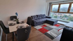 A modern living room with a dining table set for two, a grey sofa, and a large window with a city view.