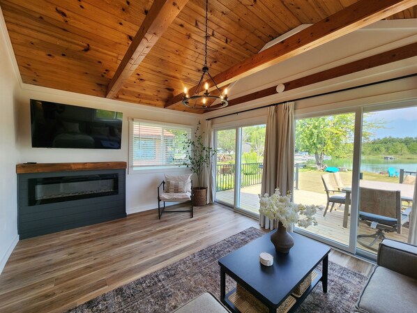 Una sala de estar moderna con chimenea, televisor y un amplio ventanal corredera que da a un área exterior con vista a un lago.