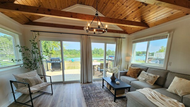 Un salón espacioso con techo de madera, ventanas amplias, un sofá gris, una mesa de centro negra y vistas a un lago y una zona de estar exterior.