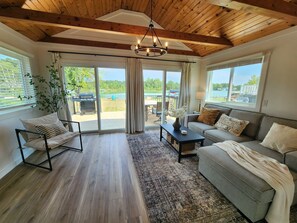 Un salón espacioso con techo de madera, ventanas amplias, un sofá gris, una mesa de centro negra y vistas a un lago y una zona de estar exterior.