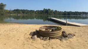 Una playa arenosa con un brasero de metal y un muelle de madera que se adentra en un lago tranquilo.