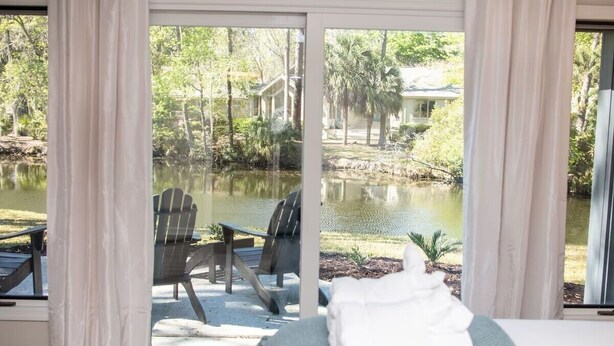 A room with a view of a lake, outdoor seating, and a towel on the bed. - Sea Pines! Beach, pool, Harbour Town Golf Links view, sleeps 8 & pet friendly! (Hilton Head Island)