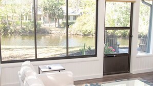 A room with a large window overlooking a pond, a white sofa, and a small table. - Sea Pines! Beach, pool, Harbour Town Golf Links view, sleeps 8 & pet friendly! (Hilton Head Island)