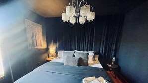 Quarto com uma cama grande, um candelabro, mesa de cabeceira com abajur e um porta-bandeja com artigos de higiene pessoal.