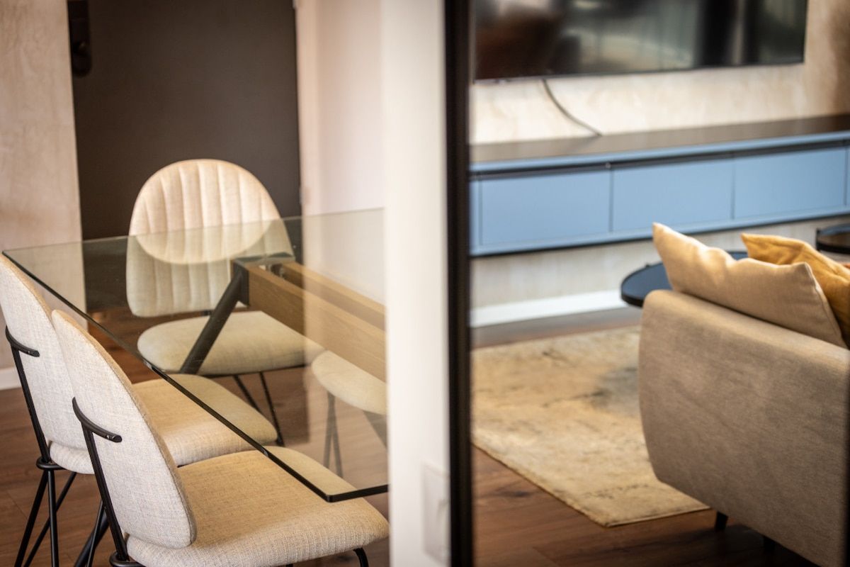 Ein modernes Interieur mit einem Glastisch, umgeben von Stühlen, einer Couch mit Kissen und Blick durch eine Glaswand.