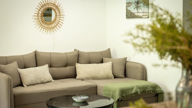 A living room with a beige sectional sofa, a round glass coffee table, a decorative mirror, and a framed picture on the wall. - Modern 2BR Apartment Near Baháʼí Gardens – Haifa (Haifa)