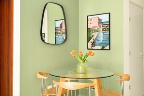 Une pièce avec une table ronde en verre, des chaises en bois, un vase de fleurs, un miroir et deux tableaux encadrés accrochés au mur.