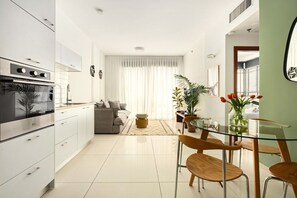 Een moderne keuken met witte kasten, een inbouwoven en een eethoek met een glazen tafel en houten stoelen.