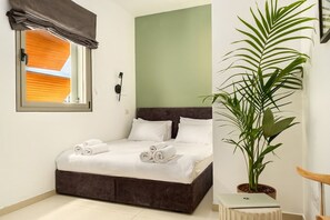 Une chambre à coucher avec un lit, une fenêtre avec des stores, une plante en pot et un ordinateur portable posé sur une table d’appoint.