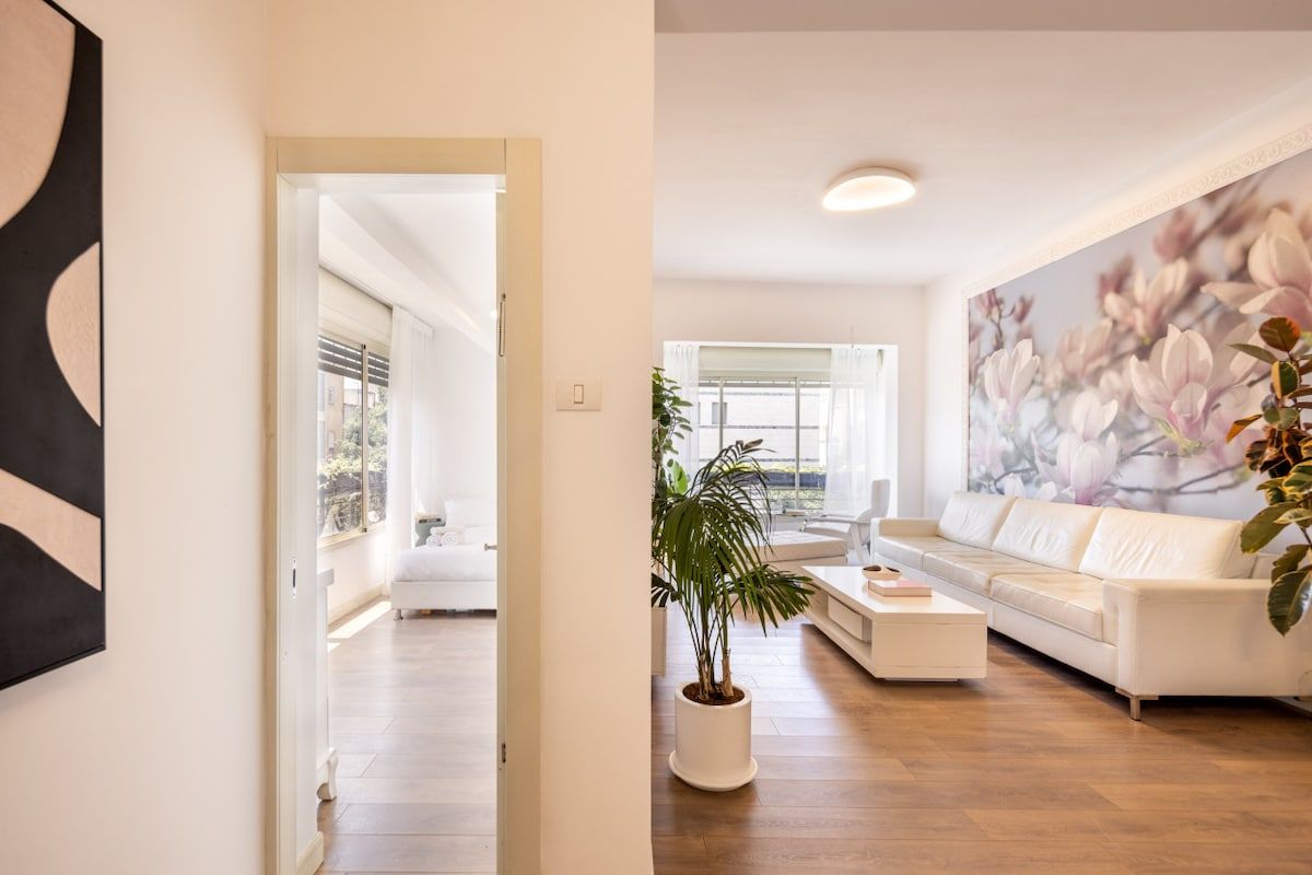 Un salón moderno con un sofá blanco, una mesita de centro y un cuadro grande con motivos florales en la pared.