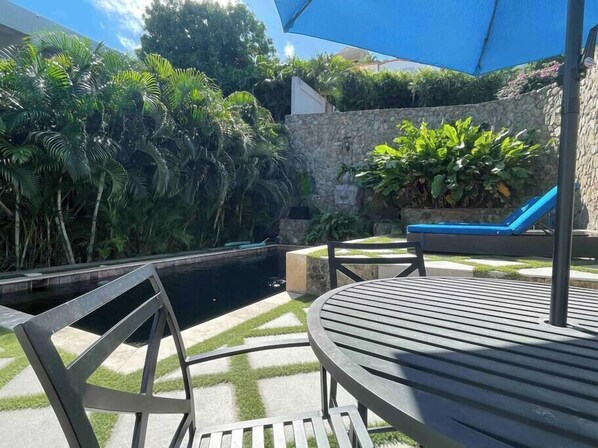 A poolside area with a stone wall, a blue lounge chair, and a table with chairs. - Garden view Simpson Bay apartment with pool-GENERATOR (2-1B) (Simpson Bay)