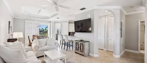 A modern living room with a white sofa, coffee table, and TV. There is a ceiling fan, a dining area with chairs, and a view of the outdoors.