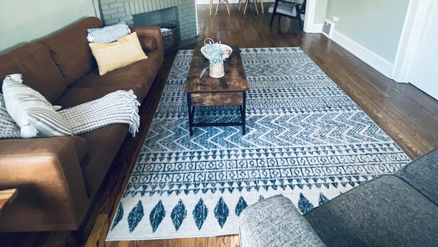 Ein Wohnzimmer mit braunem Ledersofa, einem grauen Sofa, einem Couchtisch und einem gemusterten Teppich. Im Hintergrund befindet sich ein Deckenventilator, ein Kamin und ein Essbereich.