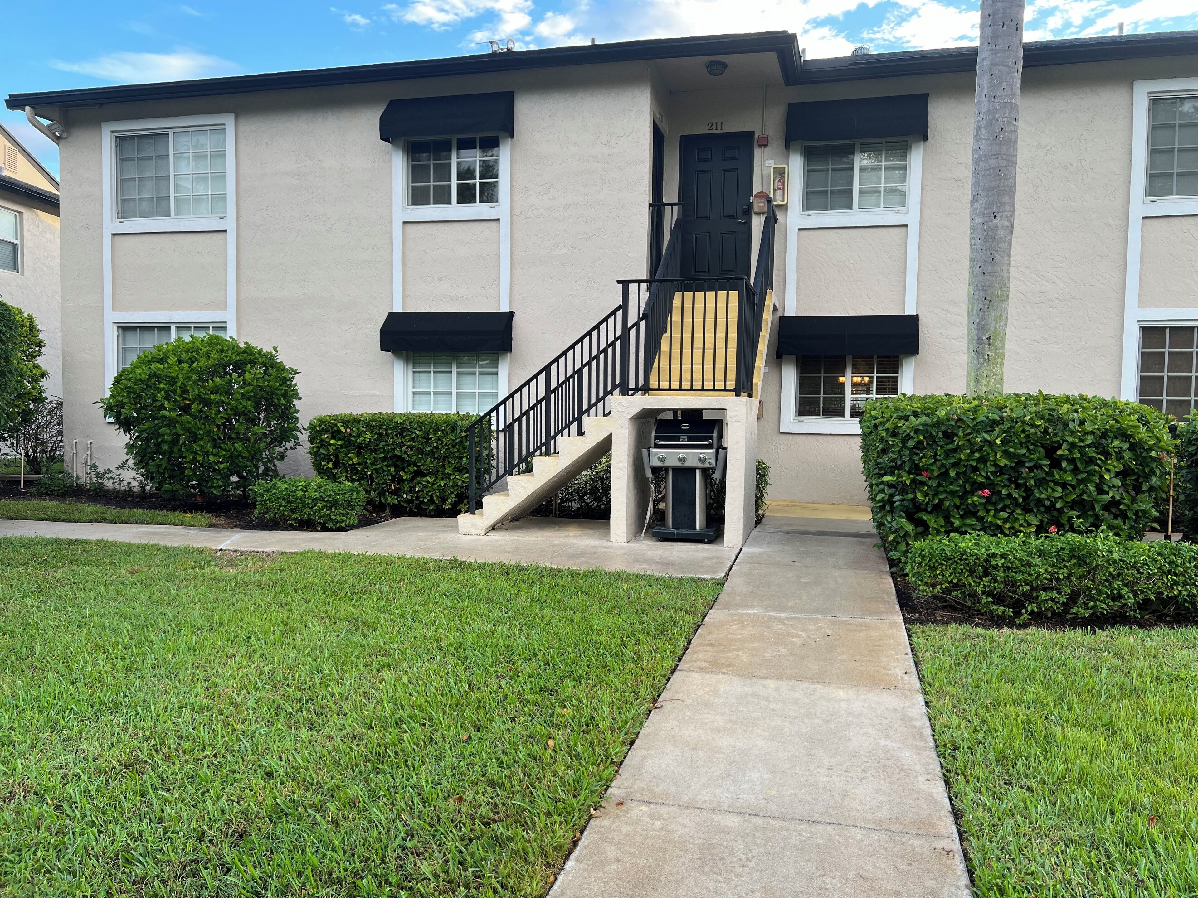 A two-story apartment building with a black front door, a small outdoor grill, and a concrete pathway leading to the entrance.