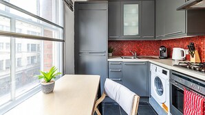 Une cuisine compacte avec des meubles gris, un lave-linge, une cuisinière et une fenêtre donnant sur un paysage urbain.