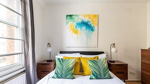Une chambre à coucher bien rangée, avec un lit, deux tables de chevet, une commode, une lampe et une œuvre d’art accrochée au mur.