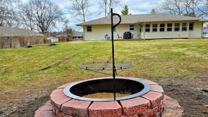 A circular brick fire pit with a metal stand in the center, surrounded by a grassy yard with a house in the background. - Rockford Cascade Getaway (Rockford)