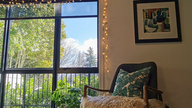 Una habitación con un ventanal grande, una silla con un plaid suave, un cuadro enmarcado en la pared y una guirnalda de luces.