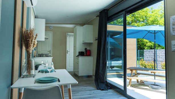Une cuisine compacte avec des meubles blancs, une table à manger dressée avec des assiettes et des verres, et une vue sur une terrasse agrémentée d’un parasol bleu.