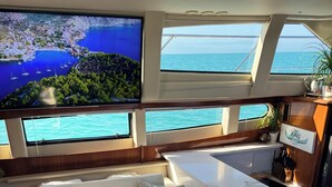 A yacht interior with a white sofa, a small kitchen area, and large windows offering ocean views.