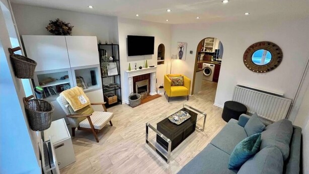 Een moderne woonkamer met een grijze bank, een klein bijzettafeltje, een gele fauteuil, een televisie en een keukenruimte zichtbaar door een boog.