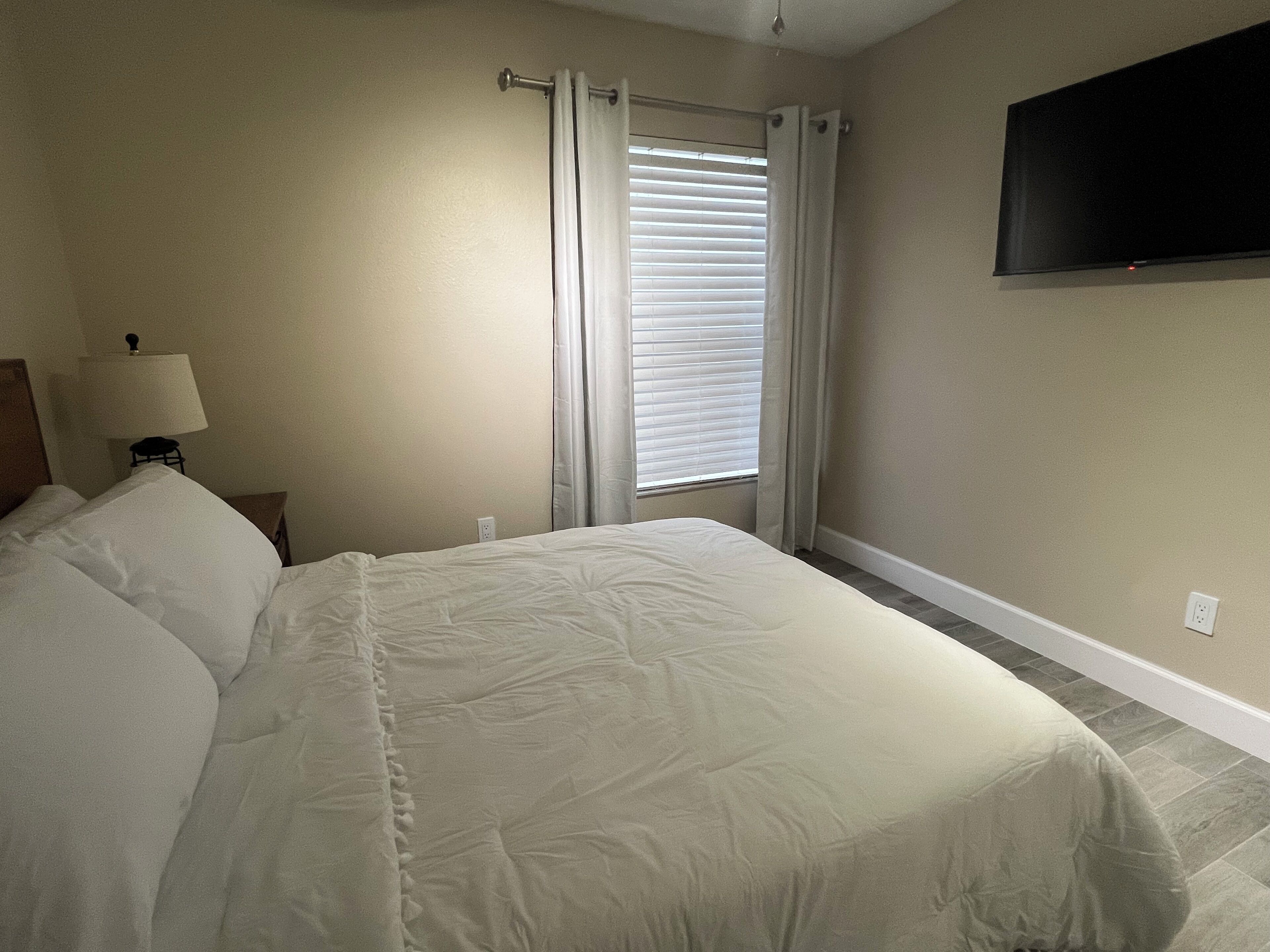 A bedroom with a large bed, a bedside lamp, a flat-screen TV mounted on the wall, and a window with blinds.