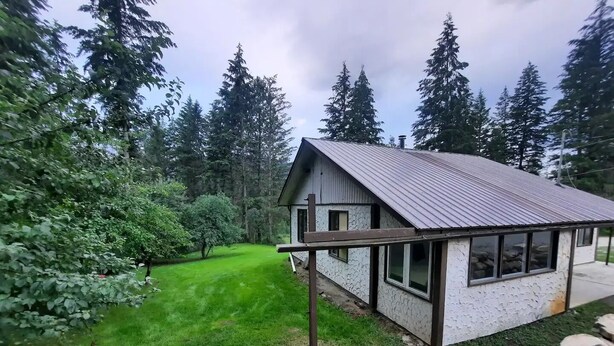 A small house with a metal roof, surrounded by a well-maintained lawn and dense trees. - Cozy 3 bedroom cottage in the woods

 (Salmon Arm)
