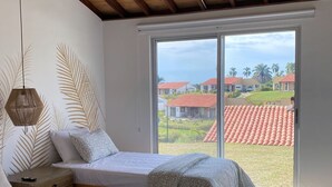 A bedroom with a large bed, a smaller bed, a ceiling fan, a window with a view of houses and palm trees, and a wooden ceiling. - Tres Colinas Estancia 27 (Armenia)