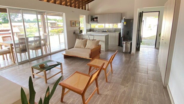 A spacious living area with a high ceiling, wooden beams, a ceiling fan, a kitchen area, and a sliding glass door leading to an outdoor patio. - Tres Colinas Estancia 27 (Armenia)