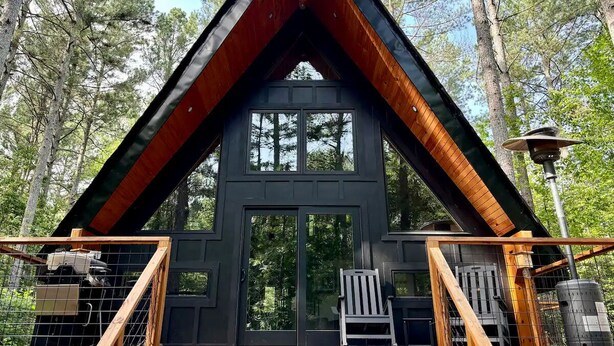 A cabin with a triangular roof, large windows, and a wooden deck with a chair.