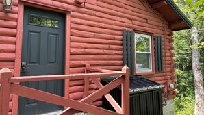Une cabane en rondins avec une porte sombre, une fenêtre avec des volets et un petit porche doté d’une balustrade.