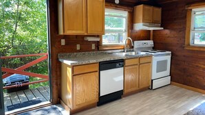Une cuisine rustique aménagée dans une cabane en bois, avec des comptoirs en granit, des appareils électroménagers blancs et une vue sur la forêt par la fenêtre.