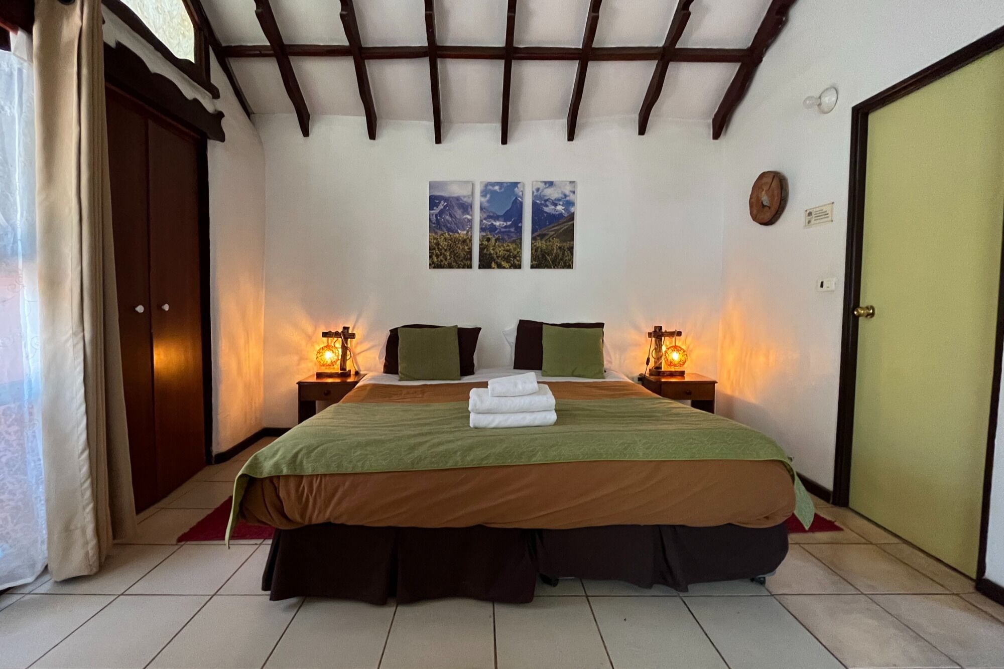A bedroom with a large bed, two bedside tables with lamps, a wooden ceiling, and a green door.