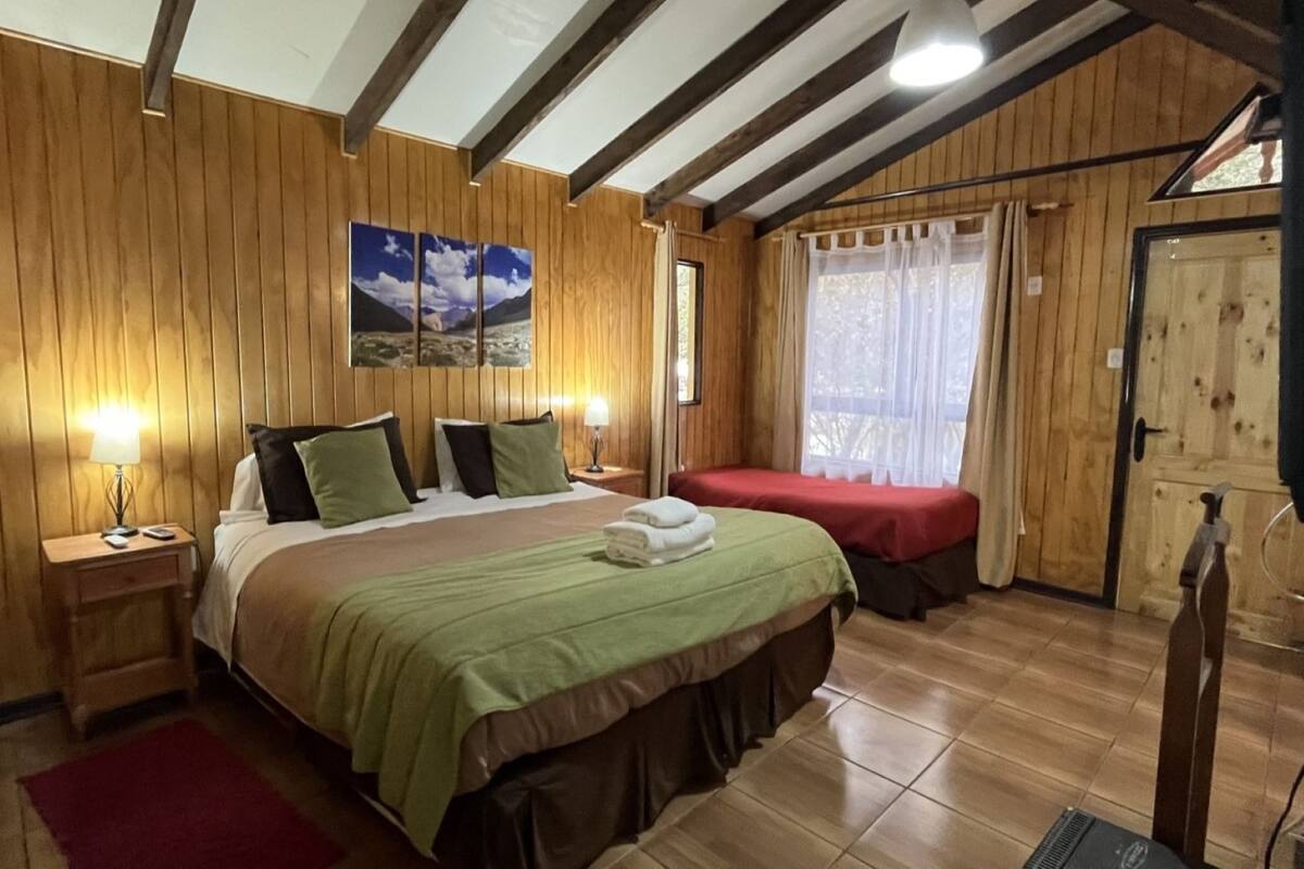 Una stanza con rivestimento in legno, un letto grande, un letto più piccolo, un comodino con una lampada e una finestra con tende.