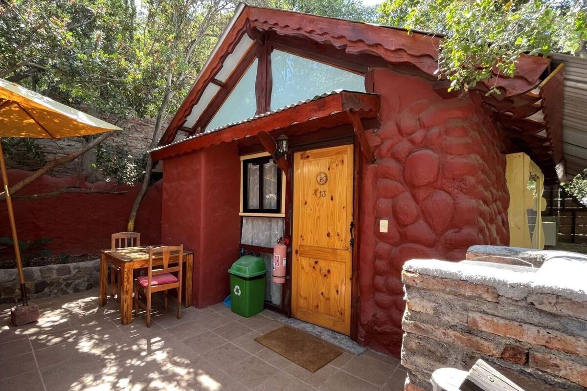Una baita di legno rustica con facciata rossa, una porta in legno con il numero 13, un tavolino con sedie e un bidone per la raccolta differenziata verde.
