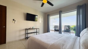 Habitación de hotel con cama, ventilador de techo, televisión, una mesita pequeña y balcón con sillas.