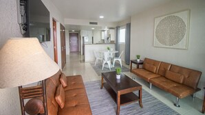 Una sala de estar moderna con un sofá marrón, una mesa de centro de madera y una lámpara. Hay un comedor con una mesa y sillas blancas, y una cocina al fondo.