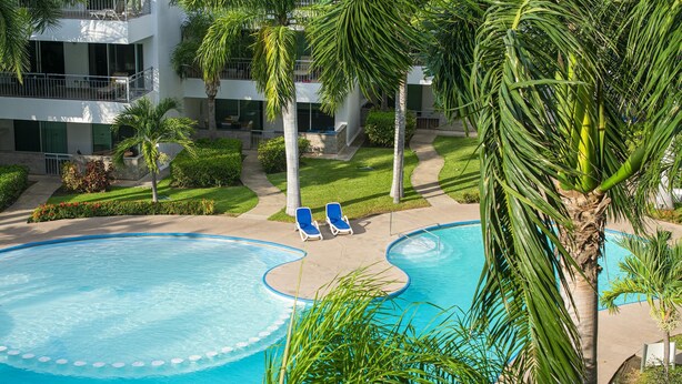 Una piscina con dos sillas reclinables azules, rodeada de palmeras y un edificio de varios pisos con balcones.