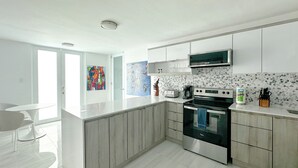 A modern kitchen with a stainless steel stove, a microwave, and a coffee maker. There is a large island with a sink and a chair. The kitchen has white cabinets and a tiled backsplash. - New Boutique House with Pool near the Beach (Rincón)
