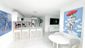 A modern kitchen with white cabinets, a central island with bar stools, a flat-screen TV mounted above the island, and a large abstract painting on the wall. - New Boutique House with Pool near the Beach (Rincón)