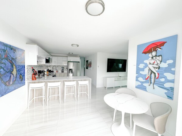 Una cocina moderna con armarios blancos, una isla central con taburetes de bar, un televisor de pantalla plana montado sobre la isla y un cuadro abstracto grande en la pared.