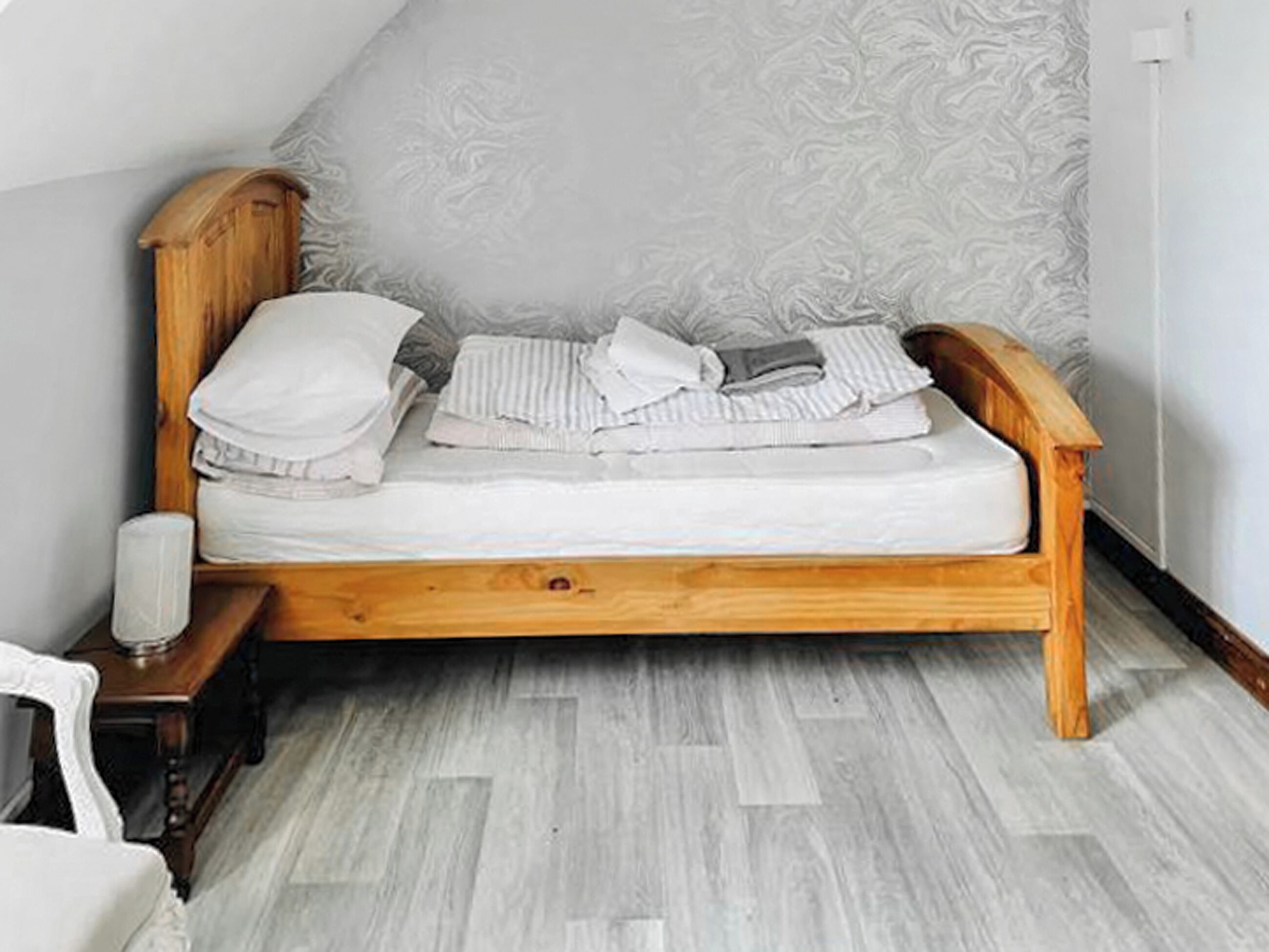 Ein hölzernes Bett mit weißer Matratze und Kissen, ein Nachttisch mit Lampe und eine Wand mit Muster.