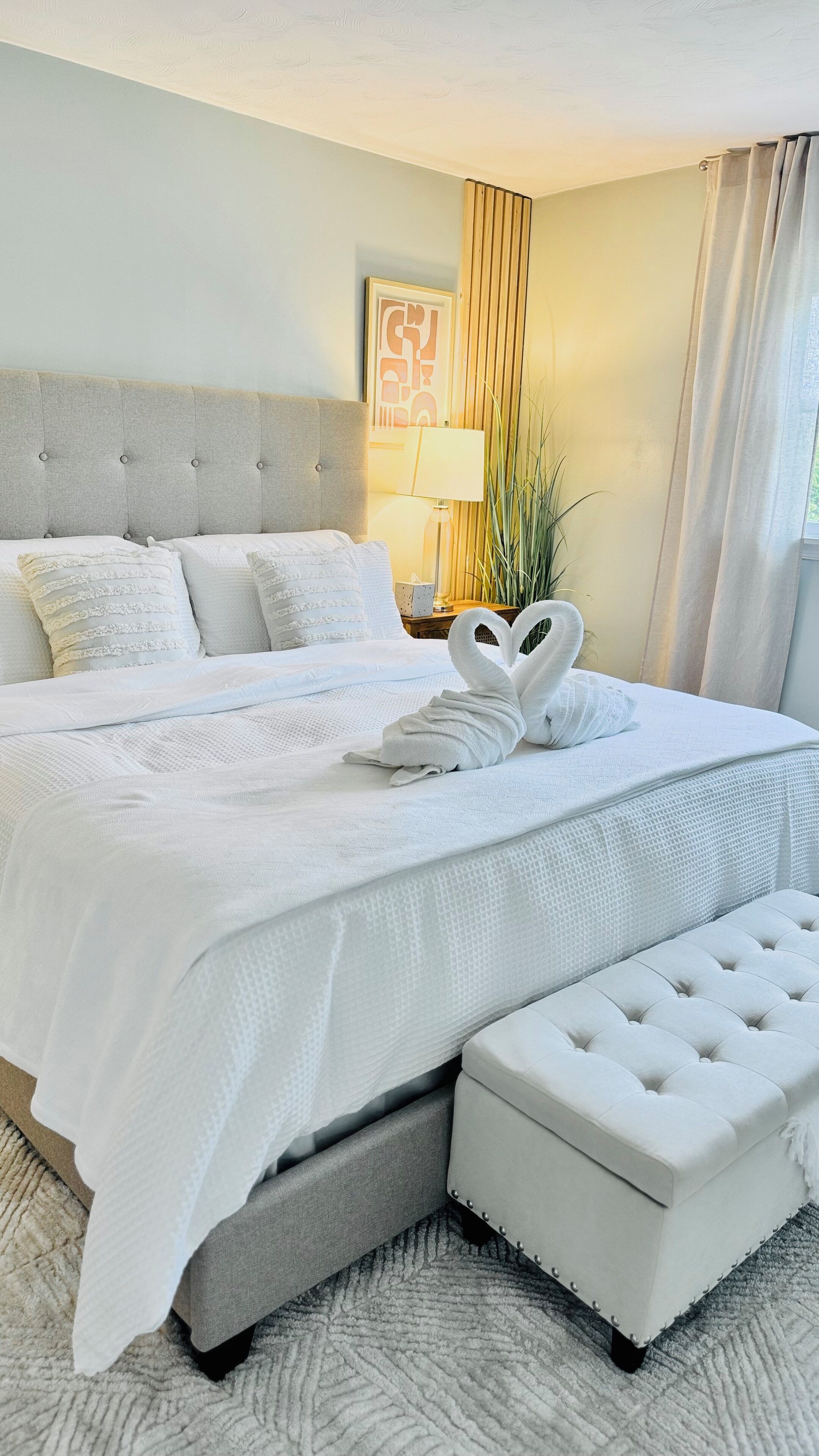 A neatly made bed with white linens and a heart-shaped towel arrangement, a tufted headboard, a bedside table with a lamp, and a window with curtains.