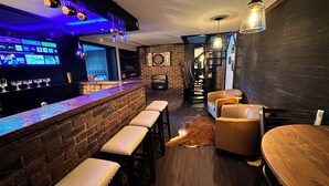 Zona de bar con mostrador de ladrillo, taburetes, sillones de cuero, un dardo y una escalera que sube a otro nivel.