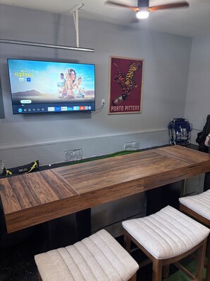 Ein moderner Essbereich mit einem langen Holztisch, gepolsterten Stühlen, einem an der Wand montierten Fernseher, der ein Programm zeigt, und einem Poster.