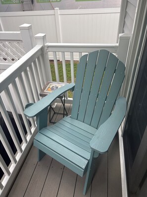 Ein blaugrüner Adirondack-Sessel steht auf einer Holzterrasse mit weißer Geländer, daneben ein kleiner Tisch mit einem dekorativen Gegenstand und im Hintergrund ein weißer Zaun.