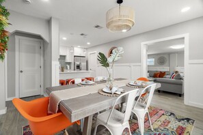 Un espace repas moderne avec une table dressée pour quatre personnes, entourée de chaises blanches et agrémentée de touches de couleur orange, avec une cuisine en arrière-plan.